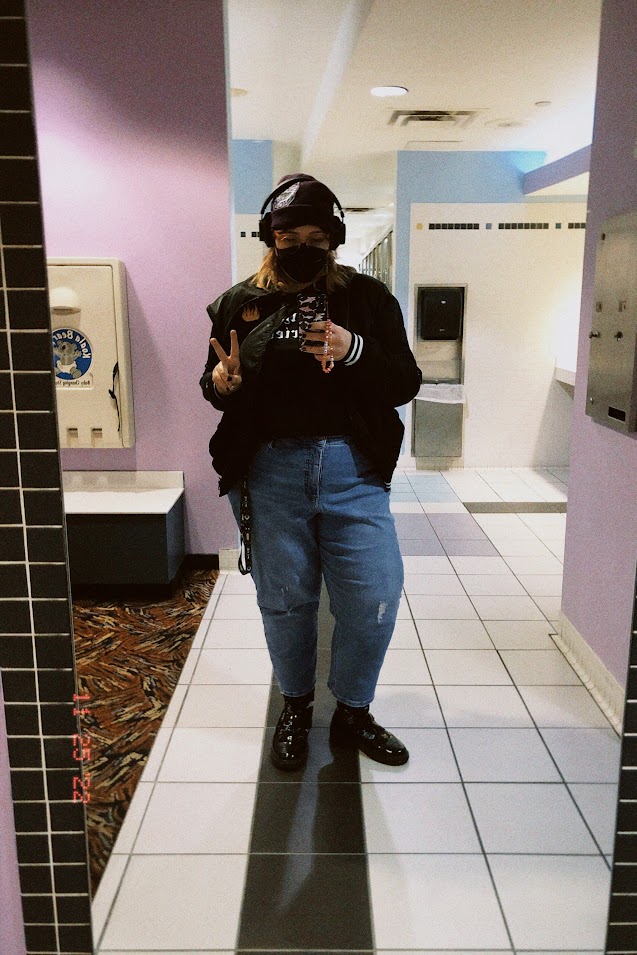 november 25, 2022: jude, a long-haired person wearing jeans, a bomber jacket with a flame on the front, and a purple beanie, takes a selfie in a full length mirror. she is making a peace sign.